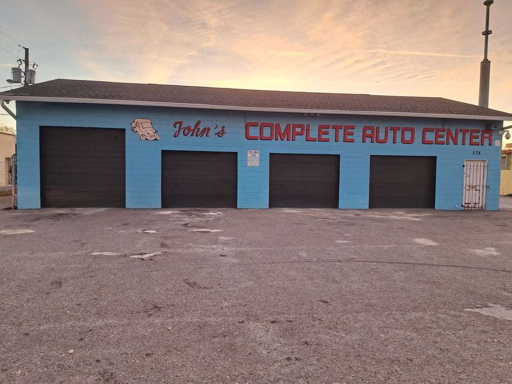 Blue business front with white cartoon car and red letters spelling john’s complete auto center, and four black garage door bays Blue business front with white cartoon car and red letters spelling john’s complete auto center, and four black garage door bays